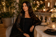 Load image into Gallery viewer, A woman seated at an outdoor dining table at night, wearing a black strapless dress with a sheer black lace shawl draped over her shoulders. The setting features warm string lights, potted plants, and tableware.